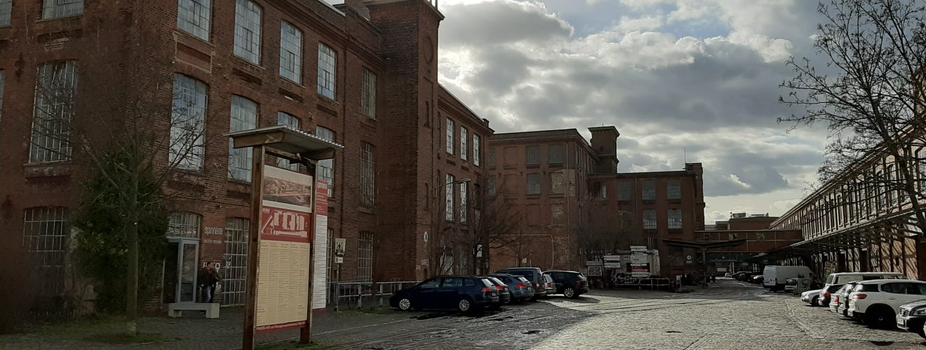 Links Gebäude der Baumwollspinnerei aus Backsteinen, rechts im Bild führt eine gepflasterte, unbefahrene Straße am Gebäude vorbei, an den Seiten parken Autos
